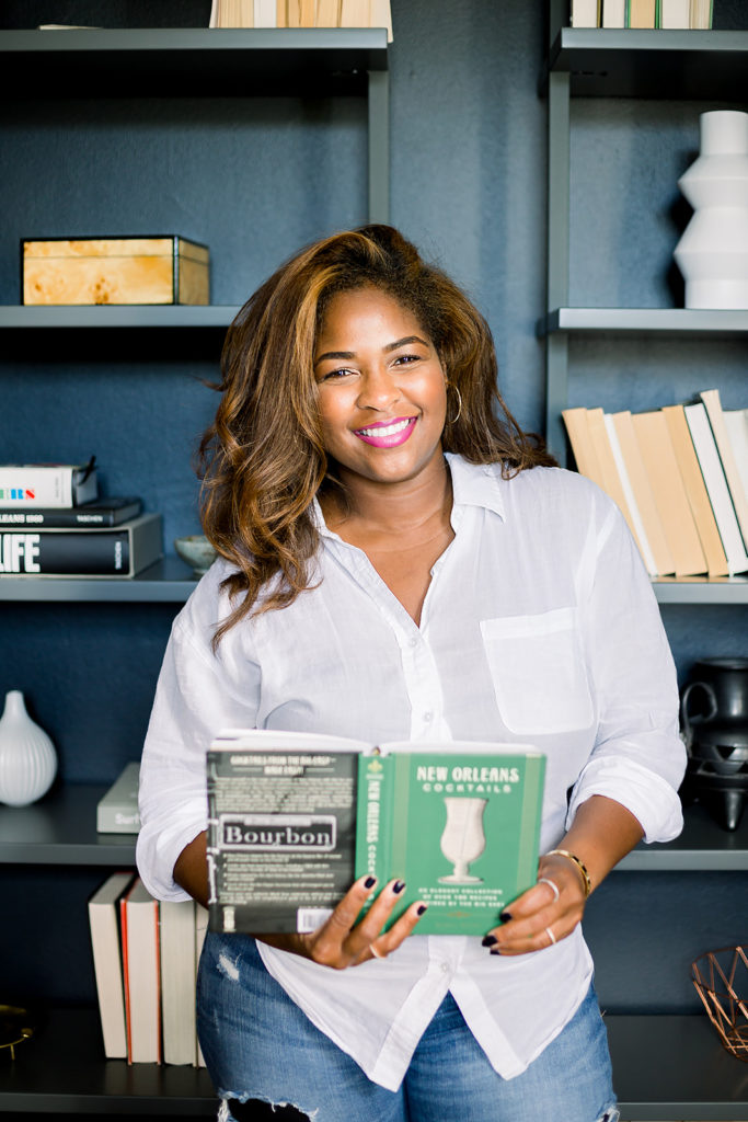 Black woman with long caramel blonde hair standing in front of book shelf holding a New Orleans cocktails book with with button down shirt and blue jeans