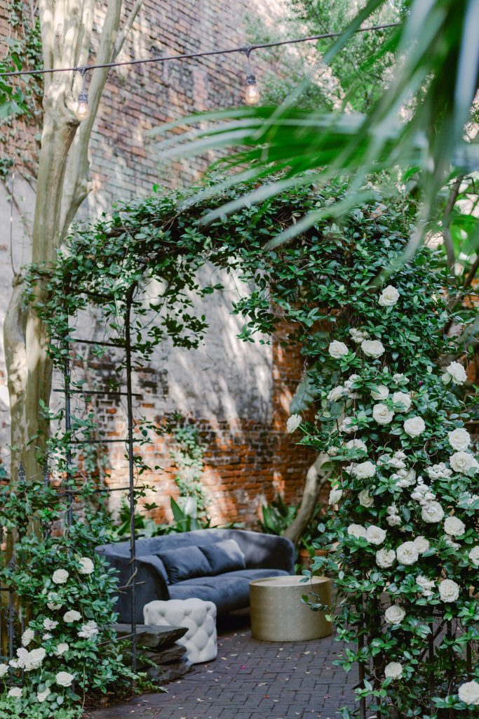 Floral arch filled with long stem white roses and dark gray sofa with white pouf and gold Coffeys table.