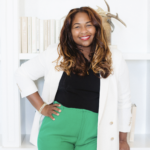 Portrait of a Black woman with long wavy hair, wearing a white jacket over a black top and green trousers, standing in a bright office space with a bookshelf and a wall-mounted antler decoration behind her.