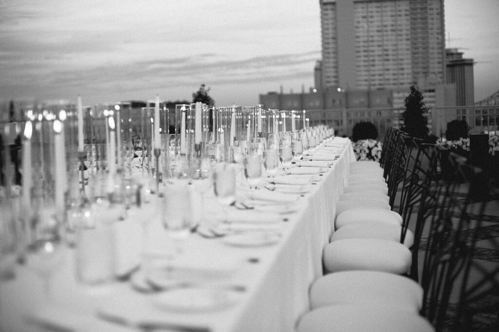 Elegant rooftop wedding table setup in black and white, with a long table lined with white chairs and glassware under a cloudy sky.