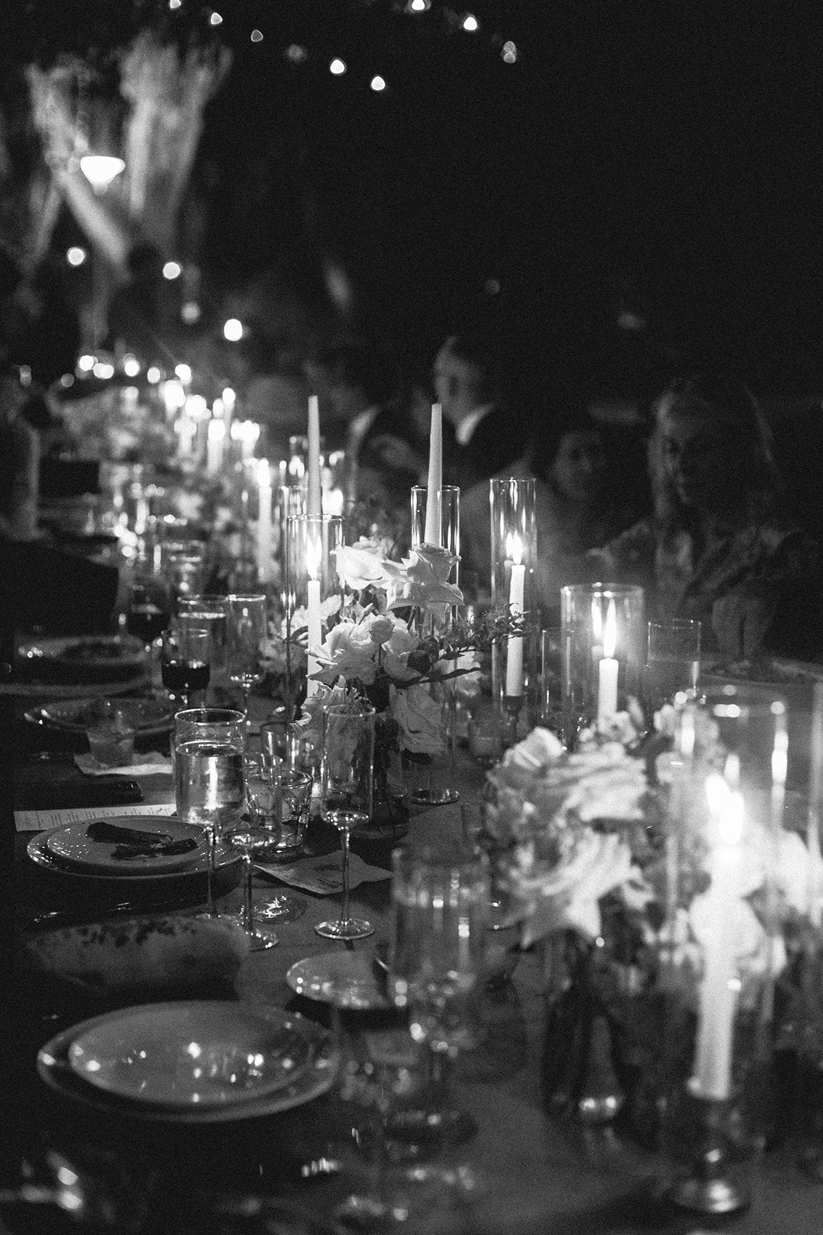 an elegant and dimly lit wedding dinner setting. The long table is adorned with numerous lit candles in tall holders, creating a warm, soft glow. The table is set with fine china and glassware, reflecting the flickering candlelight. Decorative floral arrangements add a touch of sophistication to the ambiance. The background is blurred, focusing attention on the candlelit table setting