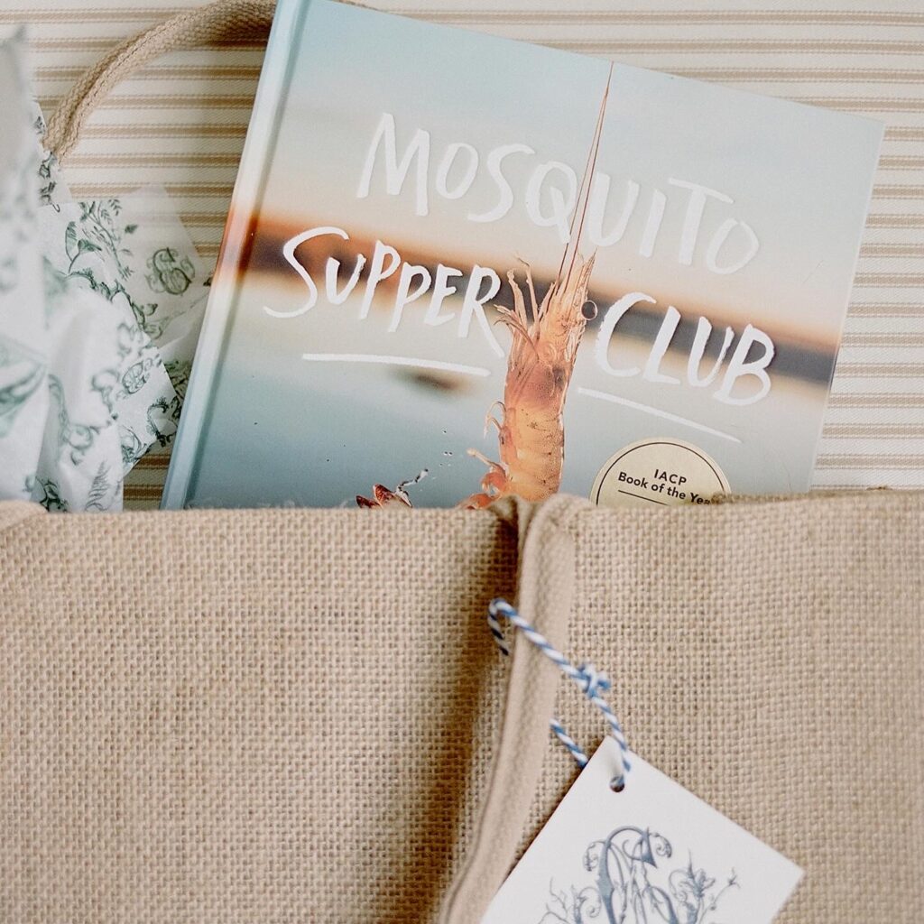 A 'Mosquito Supper Club' cookbook peeking out of a burlap welcome bag with a custom tag.